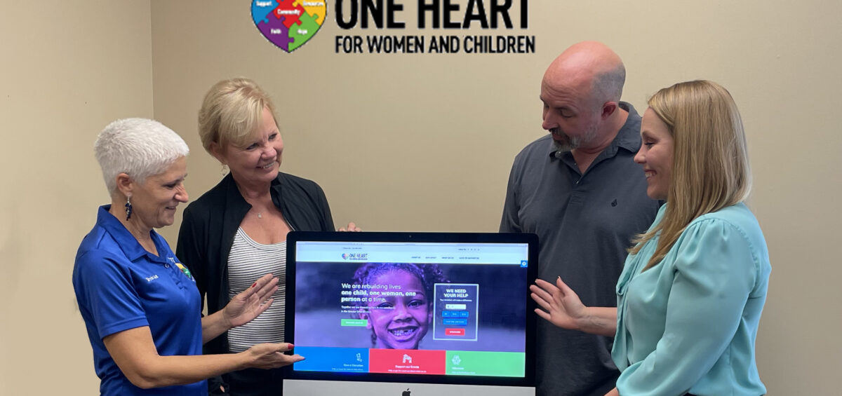 one heart for women and children founder stephanie bowman and kenney communications founder barbara kenney standon the left of a television screen displaying the new oneheartorlando.org website with frankie and maggie on the right looking down and pointing to the website