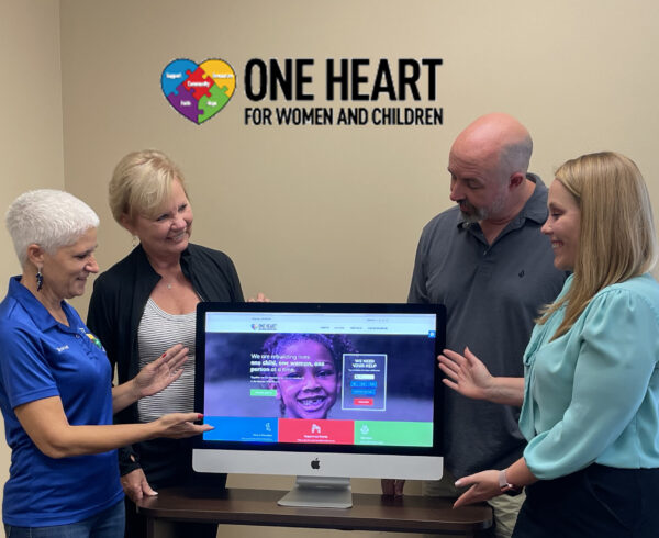 one heart for women and children founder stephanie bowman and kenney communications founder barbara kenney standon the left of a television screen displaying the new oneheartorlando.org website with frankie and maggie on the right looking down and pointing to the website